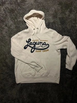 Vit hoodie från Hollister Laguna Surf - Säljer en vit hoodie från Hollister med trycket 'Laguna Surf Crew' i blått och gult på bröstet. Hoodien har huva med dragsko, känguruficka och ribbade muddar. Perfekt för en chill och avslappnad stil.