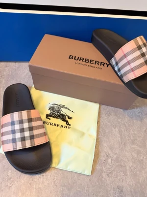 Burberry tofflor med rutigt band - Snygga tofflor från Burberry med klassiskt beige, svart och rosa rutigt band över foten. Sulan är svart och har en stilren design. Perfekta för en trendig och avslappnad look. Kommer med originalkartong och dustbag.