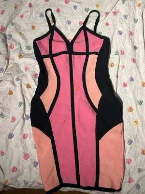 Rosa och svart bandage klänning - Säljer en kort bandage klänning i rosa och svart med tunna axelband och grafiska svarta linjer som framhäver figuren. Klänningen har v-ringning och dragkedja i ryggen. Perfekt för dig som vill sticka ut med en färgstark look.