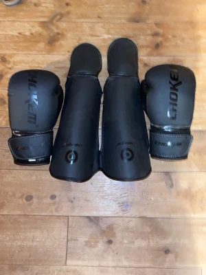 Svarta benskydd och handskar Chokem - Säljer ett set med svarta benskydd i storlek M och  14 Oz boxningshandskar från Chokem. Benskydden har kardborreband för enkel justering och handskarna har vaddering för bra skydd. Perfekt för kampsportsträning eller sparring.