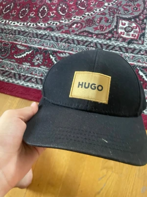 Svart keps från HUGO med logga - Snygg svart keps från HUGO med gul patch och tydlig logga framtill. Kepsen har böjd skärm och justerbar rem bak. Insidan har röda detaljer och etikett med tvättråd. Perfekt accessoar för en stilren och modern look.