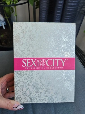 Sex and the City DVD-box - Sex and the City: The Essential Collection DVD-box med flera säsonger av serien. Boxen är silverfärgad med blommönster och har en rosa detalj med titeln. Inuti finns flera DVD-skivor med serien, snyggt förpackade i en dekorativ box.