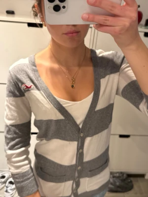 Hollister gråvit randig kofta  - Gråvit randig kofta från Hollister med breda horisontella ränder och knappstängning framtill. Storlek M passar S bättre skulle jag säga! Koftan har V-ringning, trekvartsärmar och en liten röd Hollister-logga broderad på bröstet. Perfekt att slänga över en topp för en avslappnad look.