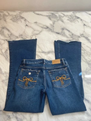 Lågmidjade bootcut jeans med snygga fickor  - Ett par blå jeans från Monki med raka ben och snygga gula rosettbroderier på bakfickorna. Klassisk femficksmodell med bälteshällor och läderpatch med Monki-logga bak i midjan. Jeansen har en trendig och ungdomlig vibe.