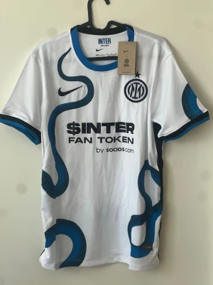 Inter Milan vit bortatröja Nike - Säljer en vit Inter Milan bortatröja från Nike med blå och svart orm-mönster, klubbmärke på bröstet och sponsortryck på framsidan. Tröjan har rund hals med blå kant och korta ärmar med blå detaljer. Perfekt för fotbollsfans!