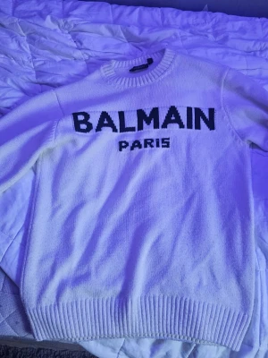 Vit stickad tröja från Balmain - Snygg vit stickad tröja från Balmain med svart text 'BALMAIN PARIS' över bröstet. Tröjan har rund halsringning och ribbade muddar vid ärmslut och nederkant. Perfekt för dig som gillar exklusiva märken och enkel, clean stil.