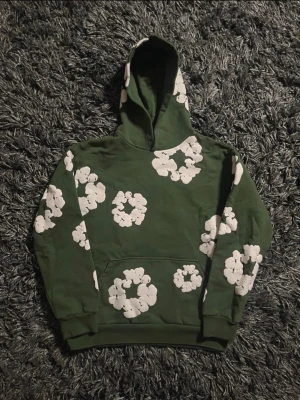 Denim tears Tröja  - Säljer en grön hoodie med stora vita blommönster över hela tröjan. Hoodien har en klassisk huva och magficka framtill. Materialet ser ut att vara mjukt och bekvämt, perfekt för en avslappnad stil. Ribbade muddar vid ärmslut och nederkant.