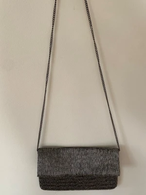Svart clutchväska med silverdetaljer - Svart clutch med tunna silverfärgade detaljer. Väskan har ett lock och en lång kedjerem så den kan bäras både i handen och över axeln🩶