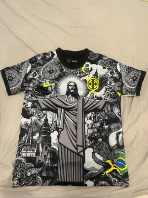 Svart Brasil t-shirt med Kristusmotiv - Unik svart t-shirt med stort tryck av Kristusstatyn från Rio de Janeiro på framsidan, detaljerade grafiska mönster och brasilianska flaggor. Korta ärmar med svartvita och gula inslag samt Brasil-logga på bröstet och ryggen. Perfekt för dig som gillar streetwear och Brasilien.