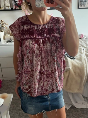 Blommig lila blus med volang - Söt blus i lila och vinröda toner med blommigt mönster och volangdetaljer längs sidorna. Blusen har rund halsringning och korta ärmar. Luftig och flowy passform.