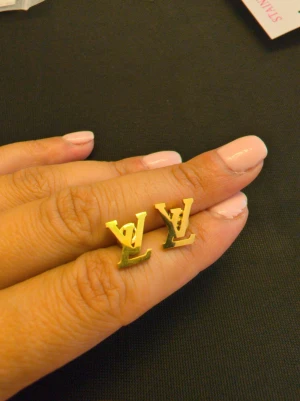 Guldiga LV örhängen i metall - Snygga örhängen i form av LV-loggan i blank guldfärgad metall. De har en stilren och modern design som verkligen sticker ut. Perfekta för dig som gillar statement-smycken och vill addera lite lyxig känsla till din look.