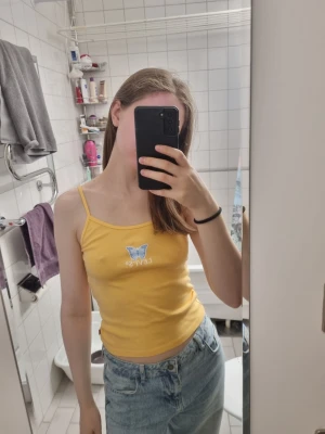 Levis Gul linnetopp med fjäril - Snygg gul linnetopp med smala axelband och en blå fjäril med texten 'Levi's' broderad framtill. Toppen är croppad och har en slim passform, perfekt för varma dagar och somriga outfits.
