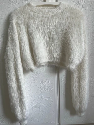 Vit fluffig cropped stickad tröja - Supermjuk och fluffig vit stickad tröja med croppad passform och långa ärmar. Tröjan har en bred ribbad rund halsringning och är gjord i ett lurvigt material som känns riktigt cozy. Perfekt för dig som gillar trendiga och bekväma plagg.