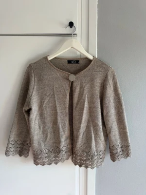 Beige stickad kofta med spetskant - Söt beige stickad kofta med rund halsringning och en dekorativ knapp framtill. Koftan har trekvartsärmar och fina spetsdetaljer längs ärmslut och nederkant. Perfekt att slänga över en topp för en chill look.