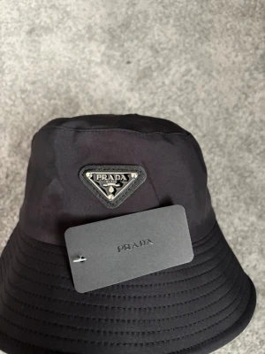 Pradabuckethat  - Snygg svart bucket hat med den ikoniska metalltriangeln framtill. Hatten är tillverkad i ett glansigt, vattenavvisande material som ger en lyxig känsla. Perfekt accessoar för att lyfta din streetwear-look.