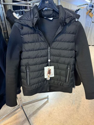 Svart pufferjacka moncler - Snygg svart pufferjacka med quiltad front och rygg, ribbade ärmar och två dragkedjeförsedda fickor. Jackan har en stor huva med dragsko och en modern, sportig look. Perfekt för kalla vinterdagar och enkel att matcha med olika outfits.