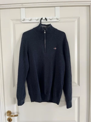 Mörkblå merinoull halfzip från Gant - Mörkblå merinoull tröja från Gant med halv dragkedja och ståkrage. Tröjan har ribbade muddar vid ärmslut och nederkant samt Gant-logga broderad på bröstet. Perfekt för lager på lager och en klassisk stil.