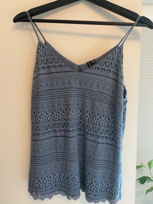 Blå spetslinnetopp från Vero Moda - Säljer en ljusblå linnetopp från Vero Moda med tunna axelband och v-ringning. Toppen är täckt av ett detaljerat spetsmönster och har en rak passform. Perfekt för sommaren och enkel att styla med jeans eller kjol.