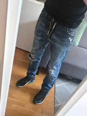 Blå slitna jeans med hål - Säljer ett par blå orginal dsq2 jeans med slitningar, hål och färgstänk för en riktigt edgy look. Jeansen har rak passform och klassisk femficksdesign. Perfekta för dig som gillar streetstyle och vill sticka ut lite extra.