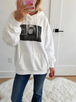Vit hoodie med tryck och huva - Säljer en vit hoodie med svartvit grafiskt tryck på bröstet och stor känguruficka framtill. Hoodien har dragsko i huvan och ribbade muddar vid ärmar och nederkant. Perfekt för en chill och avslappnad stil.