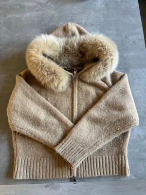 Beige stickad jacka med pälskant från Alessa  - Säljer en beige stickad jacka med dragkedja och stor huva kantad med mjuk, fluffig päls. Jackan har ribbade muddar och nederkant för extra komfort. Perfekt för kyliga dagar när du vill vara både varm och snygg.