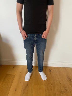 Nudie Jeans Skinny Lin - Säljer ett par blå slim jeans med klassisk femficksdesign och snygga slitningar. Jeansen har normal midja och smal passform som sitter tajt längs benen. Perfekta för en avslappnad och trendig stil. Jeansen passar även till lite större storlek en vad jeansen ursprungliga storlek