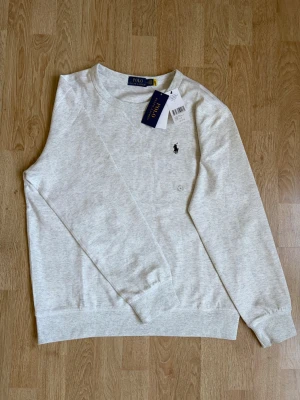 Ljusgrå sweatshirt från Polo Ralph Lauren - Ljusgrå sweatshirt från Polo Ralph Lauren med klassisk rund halsringning och broderad logotyp på bröstet. Tröjan har ribbade muddar vid ärmslut och nederkant, och är tillverkad i mjukt bomullsmaterial för en skön känsla. Tröjan är helt ny och oanvänd, skriv gärna vid frågor eller funderingar!