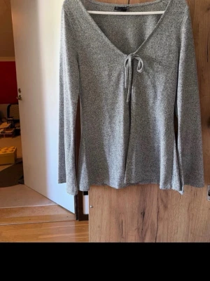 Grå långärmad topp med knyt - Säljer en grå långärmad topp med v-ringning och knyt framtill. Toppen har en mjuk och stretchig känsla, perfekt till jeans eller kjol. Enkel men ändå snygg design som passar till många olika stilar.
