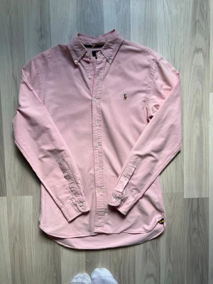 Rosa skjorta från Polo Ralph Lauren - Säljer en ljusrosa skjorta från Polo Ralph Lauren i slim fit-modell. Skjortan har klassisk krage med knappar, broderad logga på bröstet och långa ärmar. Tillverkad i mjuk bomull och perfekt för en clean och stilren look. Lite defekter se bilder 