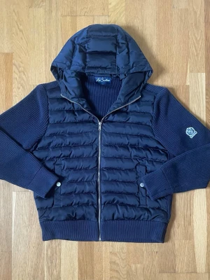 La Santino Quiltad Cardigan med Luva – Marinblå | Strl M - Stilren marinblå La Santino cardigan med quiltad framsida och huva. Kombinationen av den vadderade framsidan och de stickade ärmarna ger både värme och en exklusiv look. Passar perfekt till både vardag och finare tillfällen.  ✔ Mycket fint skick ✔ Storlek M ✔ Marinblå färg ✔ Quiltad framsida med huva ✔ Stickade ärmar och rygg ✔ Mjuk och bekväm kvalitet Hör gärna av dig om du har några frågor eller vill ha fler bilder. Jag är alltid öppen för bud! 😊