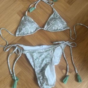 Grön och vit bikini med knyt - Snygg bikini i vitt och ljusgrönt med paisleymönster. Både överdel och underdel har knytband med gröna tofsar. Triangelformad topp och klassisk bikiniunderdel