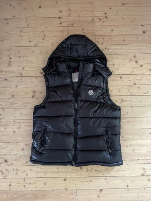 Moncler dunväst  - Snygg svart dunväst från Moncler med glansig finish och vadderad design. Västen har en praktisk huva, två dragkedjeförsedda fickor och Moncler-logga på bröstet. Perfekt för lager-på-lager och håller dig varm under kyliga dagar.