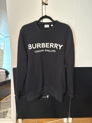 Svart hoodie från Burberry - Svart hoodie från Burberry med vit logga och texten 'London England' på bröstet. Klassisk modell med rund hals och långa ärmar. Perfekt för en clean och stilren look.