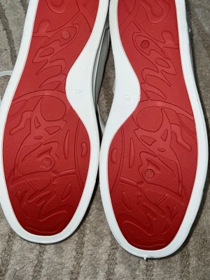 Vita sneakers med röda sulor - Snygga vita sneakers med röda yttersulor och beige/brun vadderad kant runt öppningen. Skorna har klassisk snörning och ovandelen verkar vara i syntetmaterial eller läderimitation. Sulan har ett unikt mönster i rött som sticker ut.