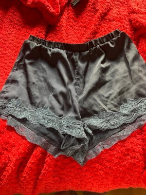 Två par pyjamasshorts i olika stilar - Säljer två par pyjamasshorts. Ett par är svarta i satin med spetsdetaljer längs benen och elastisk midja. Det andra paret är vita med små mönster, mjukt bomullstyg och dragsko i midjan. Perfekta för mysiga kvällar hemma.