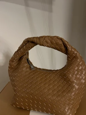 Brun flätad handväska i konstläder - Snygg brun flätad handväska använd max två gånger. Inga defekter eller fläckar på varken utsidan eller innuti. Man får plats med hur mycket som helst samtidigt som den är liten, finns även ett innerfack. Köptes på Gina för 399