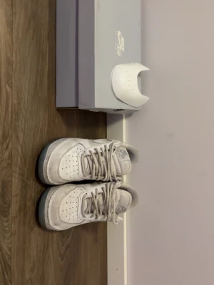 Vita Nike Air Force 1 sneakers - Klassiska vita Nike Air Force 1 sneakers med rund tå och snörning. Skorna har en platt sula och är tillverkade i skinn med perforerade detaljer på tån för extra ventilation. Perfekta för dig som gillar en clean och tidlös stil.