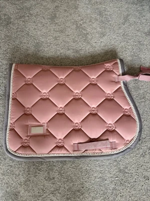 Rosa schabrak från Equestrian Stockholm - Säljer ett snyggt rosa schabrak från ES i storlek ponny, med quiltat mönster och grå kant med pärlor. Schabraket har dekorativa detaljer och broderade mönster i rutnät, samt vita pärlor längs kanten. Säljer det eftersom att den inte kommer till andvändning längre.