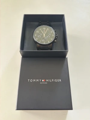 Svart klocka från Tommy Hilfiger - Stilren svart klocka från Tommy Hilfiger med metallänk och rund urtavla. Klockan har flera små urtavlor och tydliga siffror i vitt. Kommer i originalask med logga. Perfekt accessoar för dig som gillar klassisk och modern stil. Klockan är helt ny och är använd 1 gång. Det finns inga tydliga tecken på repor eller slitage. Mitt pris startar på 2300kr, priset kan diskuteras. (Endast seriösa köpare)