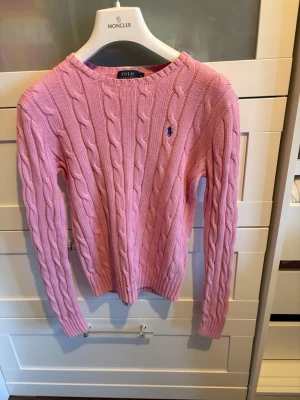 Rosa kabelstickad tröja från Polo Ralph Lauren - Snygg rosa kabelstickad tröja från Polo Ralph Lauren med rund halsringning och broderad logga på bröstet. Tröjan har långa ärmar och ribbade muddar vid ärmslut och nederkant. Perfekt för en trendig och avslappnad look. Storlek s, passar även xs