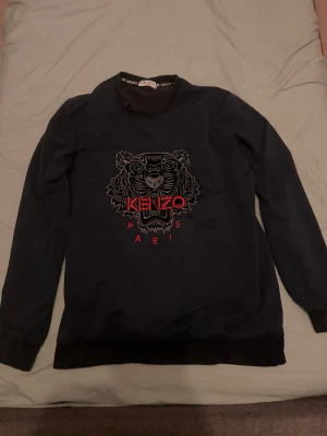 Svart stickad Kenzo tröja med tiger - Snygg svart stickad tröja från Kenzo med det ikoniska tigermotivet broderat framtill och röda detaljer. Tröjan har rund halsringning, långa ärmar och ribbade muddar vid ärmslut och nederkant. Perfekt för dig som gillar streetwear och vill sticka ut.