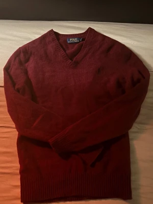 Polo Ralph Lauren Stickad Tröja - Vinröd stickad tröja från Polo Ralph Lauren med V-ringning och diskret broderad logga på bröstet. Tröjan har ribbade muddar vid ärmslut och nederkant. Perfekt för dig som gillar klassisk och stilren look.