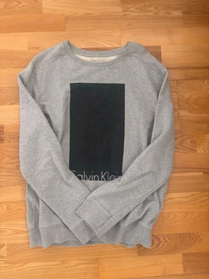 Grå stickad tröja Calvin Klein - Snygg grå stickad tröja från Calvin Klein med ett stort svart rektangulärt tryck på bröstet och diskret logga längst ner. Tröjan har rund halsringning och ribbade muddar vid ärmslut och nederkant. Perfekt för en clean och stilren look.