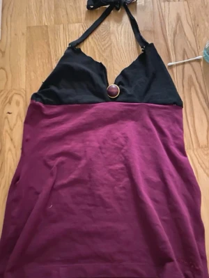 Lila och svart halterneck topp - Snygg halterneck topp i lila och svart med dekorativ ring framtill. Toppen har knyt i nacken och är ärmlös, perfekt för varma dagar eller som ett statement-plagg. Materialet känns mjukt och stretchigt. Jag har sytt den själv men jag tror att den skulle vara en S eller Xs.