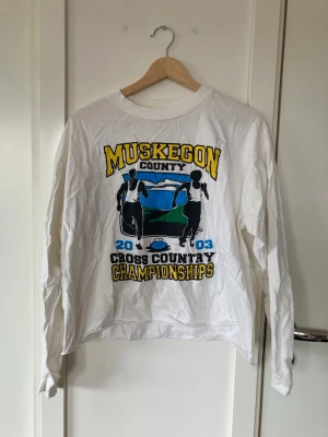 Vit vintage långärmad Muskegon t-shirt - Säljer en vit långärmad t-shirt med tryck från Muskegon County Cross Country Championships 2003. T-shirten har färgglatt motiv med löpare och text i gult, svart och blått. Perfekt för dig som gillar sportiga vintageplagg. Liten fläck på bild två på framsidan. Avklippt på längden för lite mer cropped fit. 