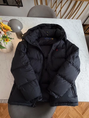Svart pufferjacka med huva - Snygg svart pufferjacka från Polo Ralph Lauren med huva och dragkedja framtill. Jackan har två sidofickor med dragkedja och diskret röd logga på bröstet. Tillverkad i ett glansigt material som ger en modern look och håller dig varm under kalla dagar.Strorel S men passar också som M