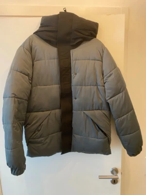 Grå pufferjacka med svart huva - Snygg grå pufferjacka med svart huva och dragkedja framtill. Jackan har två snedställda fickor med dragkedja och bred quiltad design. Perfekt för kalla vinterdagar och har en modern, oversized look.