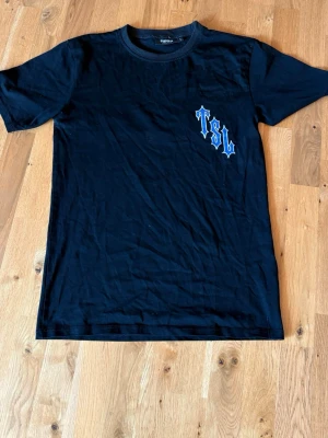 Trapstar tshirt - Svart T-shirt från Trapstar med blått och vitt TSL-tryck på bröstet. Klassisk rund hals och korta ärmar. Tillverkad i mjuk bomull som är skön att bära. Perfekt för en streetwear-look.