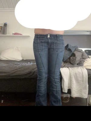 Blå utsvängda jeans med fickor - Snygga blå jeans med utsvängda ben och markerade sömmar. Jeansen har två stora fickor fram, dubbla knappar i midjan och klassiska bakfickor. Perfekta för dig som gillar retrostil och vill ha ett par jeans med coola detaljer. Har en lagning fram vid gylfen. 