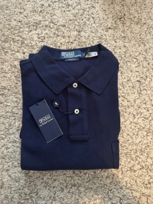 Marinblå pikétröja från Ralph Lauren - Klassisk marinblå pikétröja från Polo Ralph Lauren i modellen The Big Fit. Tröjan har krage, knappar framtill och är tillverkad i bomull. Perfekt för en stilren och avslappnad look.